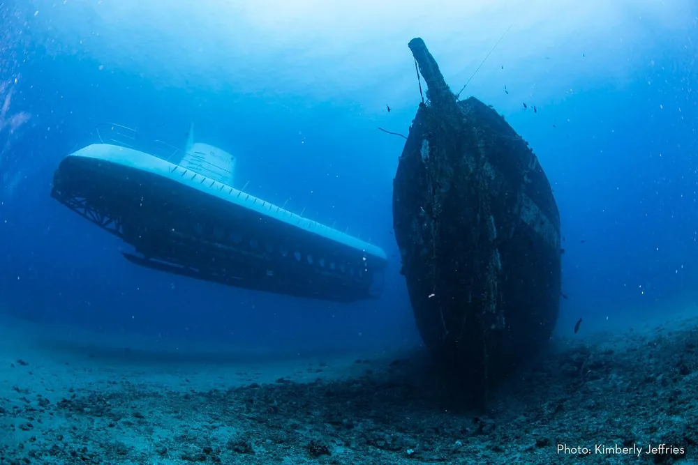 A white submarine beside the hull of a sunken ship on a sandy sea floor