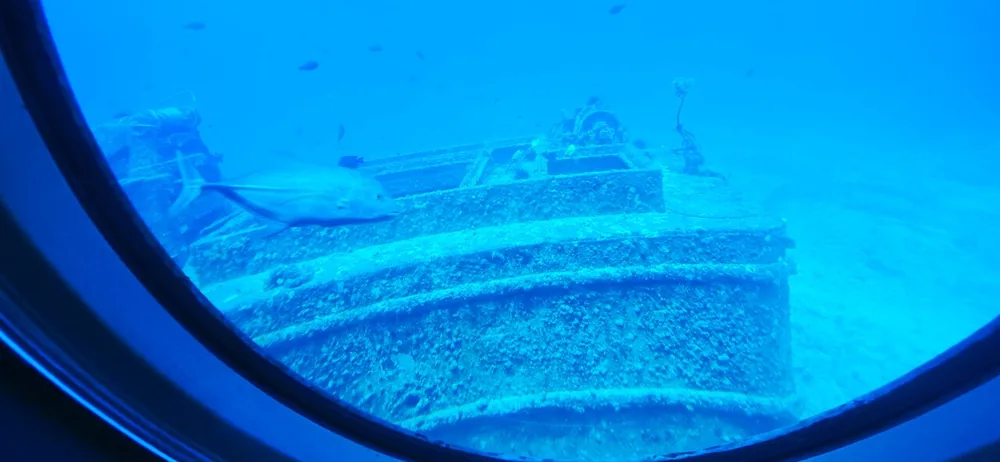 Guest photo through a round porthole of a wreck lying on the sand in blue water