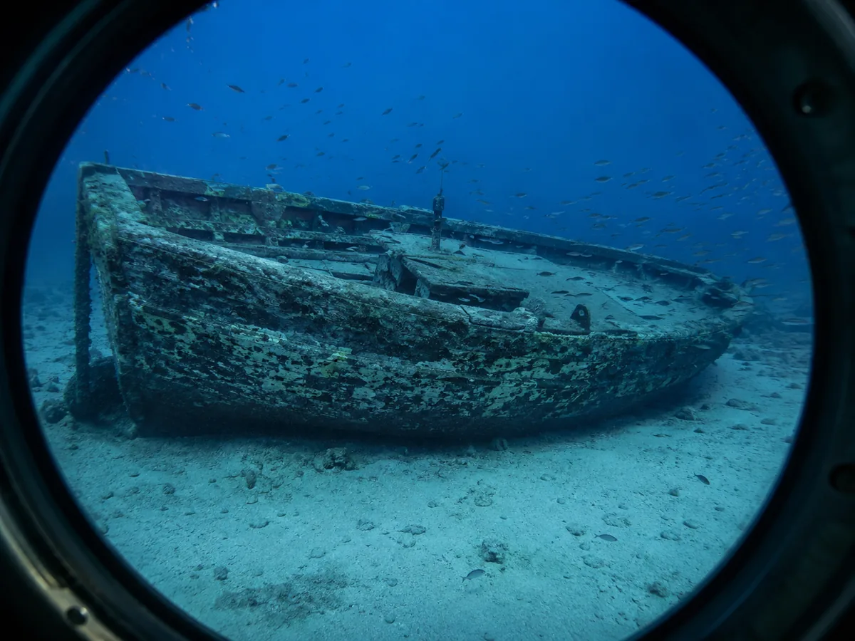 A small sunken wooden boat on white sand seen through a round submarine porthole